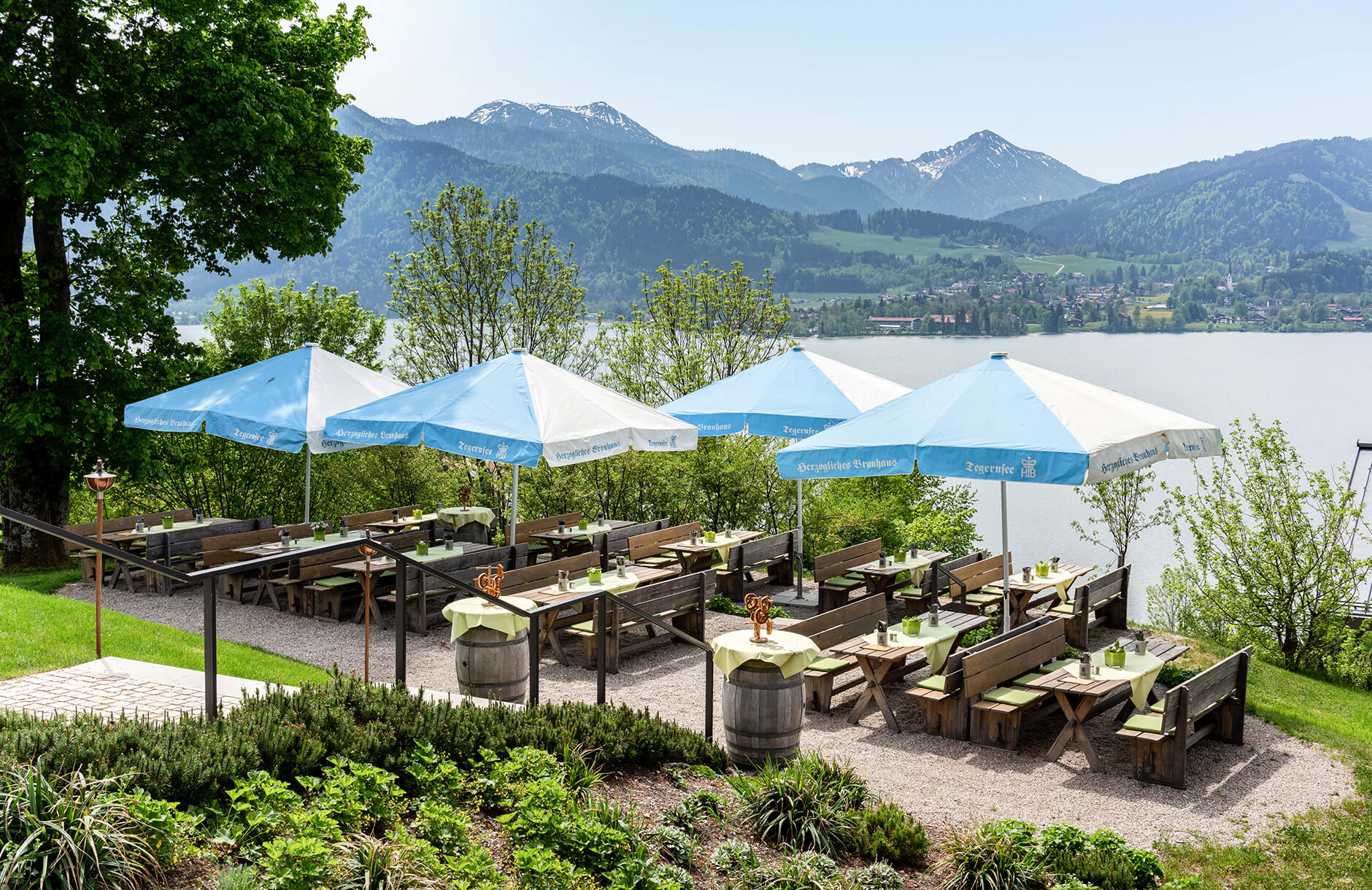 Blick auf den Biergarten auf der Terrasse mit Holzgarnituren und blau-weißen Schirmen, umgeben von Wiese und Bäumen mit Blick auf den Tegernsee und die Berge im Hintergrund.