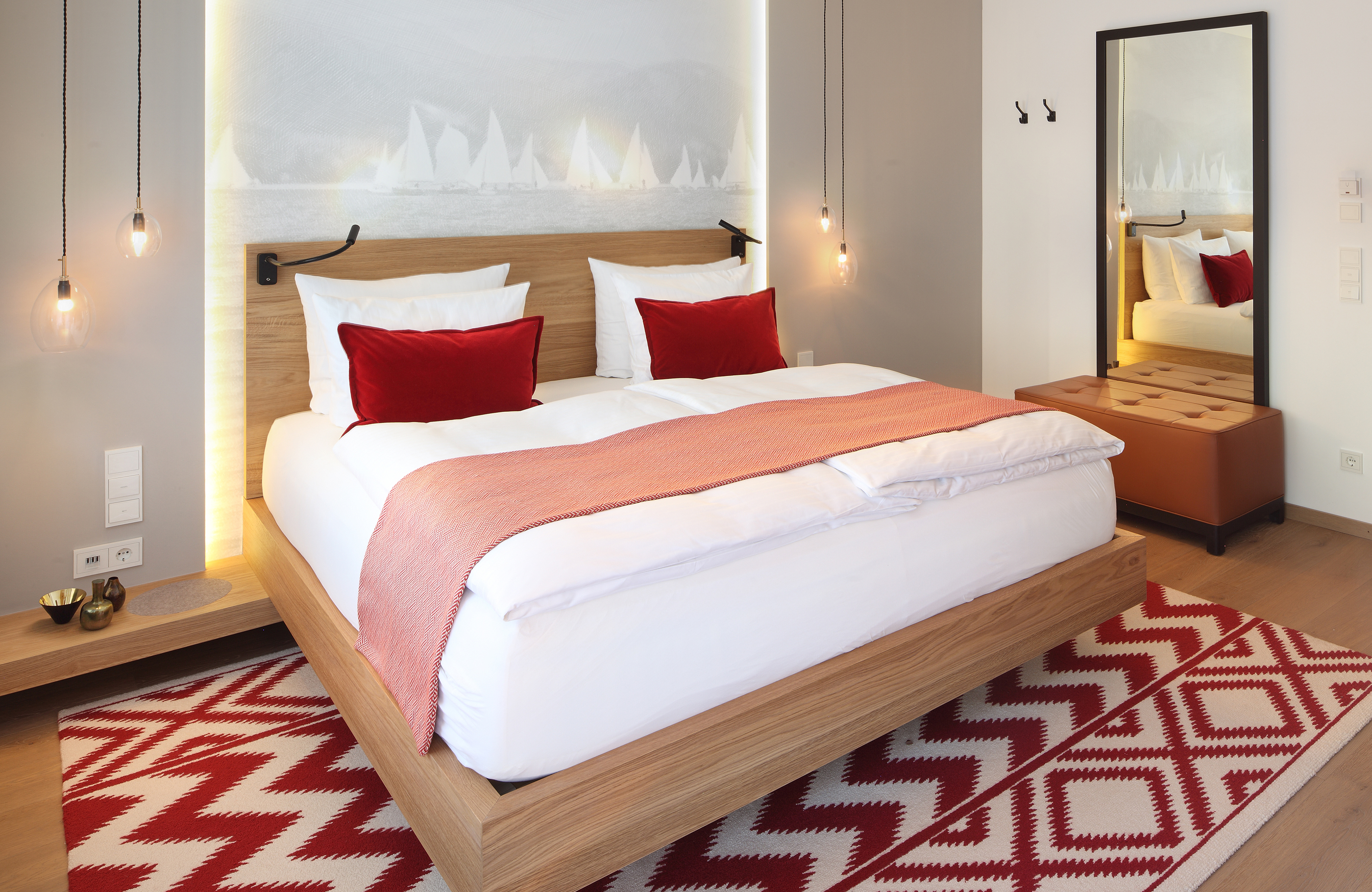 Aufnahme eines Doppelzimmers im Haus Tegernsee im Hotel Das Tegernsee mit einem großen, weiß bezogenem Doppelbett mit roten Dekokissen in der Raummitte und einem  weiß-roten Teppich.